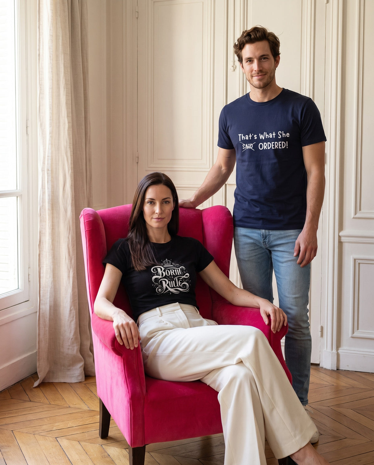 A woman in a black Born to Rule tee sits in a hot-pink velvet armchair; a man in a navy That's What She Ordered tee stands behind her with a hand on the chair.